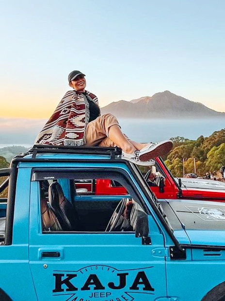 Person sitting on a blue jeep with Mount Batur in the background at sunrise, Bali.