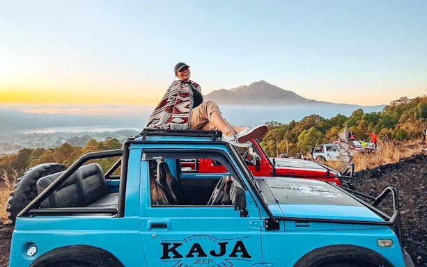 Person sitting on a blue jeep with Mount Batur in the background at sunrise, Bali.