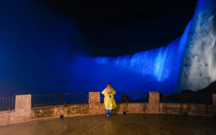Person in yellow raincoat viewing illuminated Niagara Falls at night.