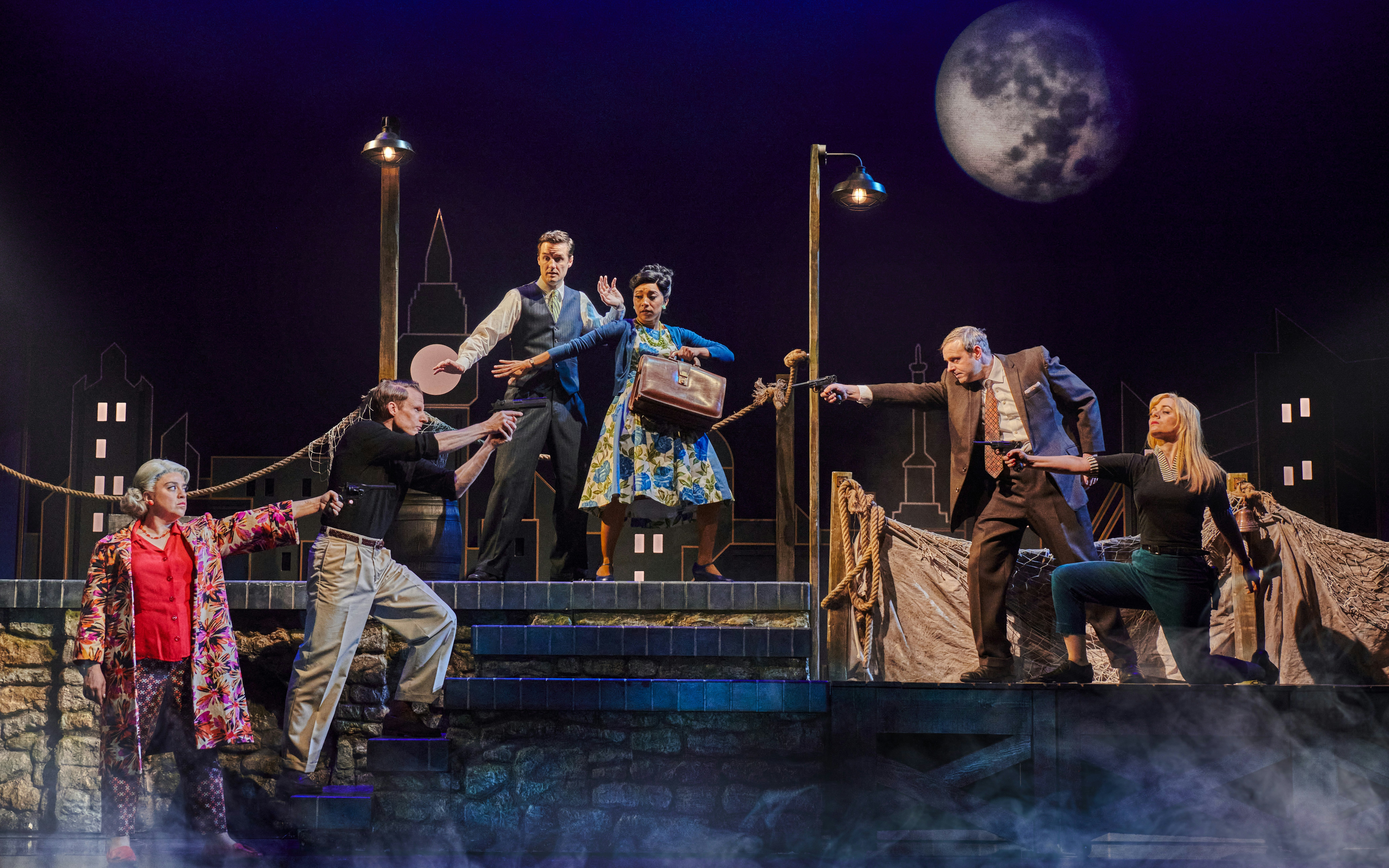 Actors performing a dramatic scene in "The Comedy About Spies" with a cityscape backdrop.