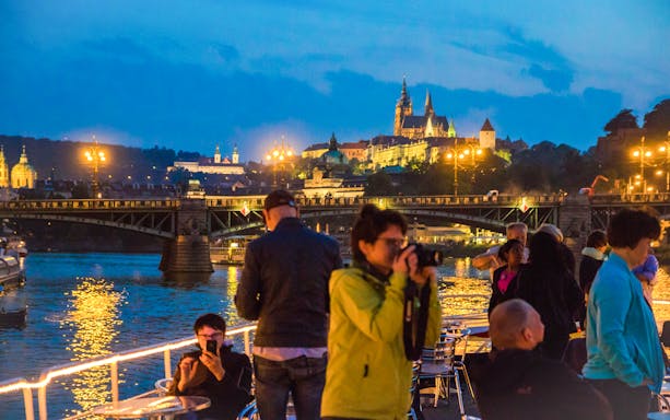 Tourists on a boat with Prague Castle and Charles Bridge illuminated at night.