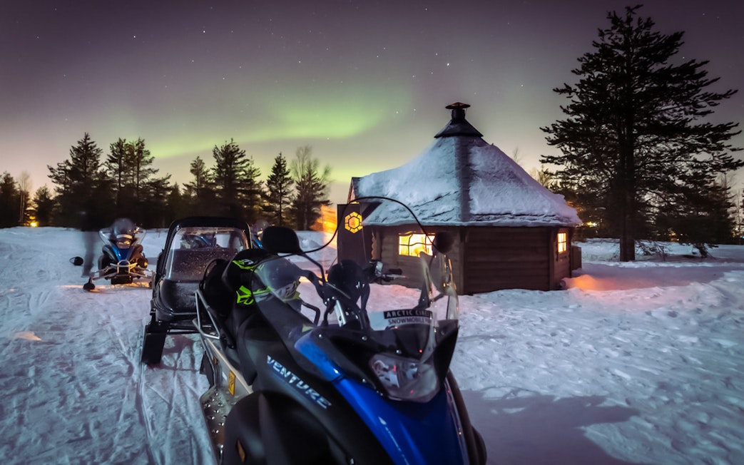 Snowmobiles parked near a Kota hut under Northern Lights in Levi, Finland.