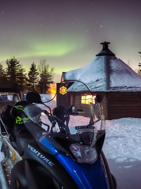 Snowmobiles parked near a Kota hut under Northern Lights in Levi, Finland.