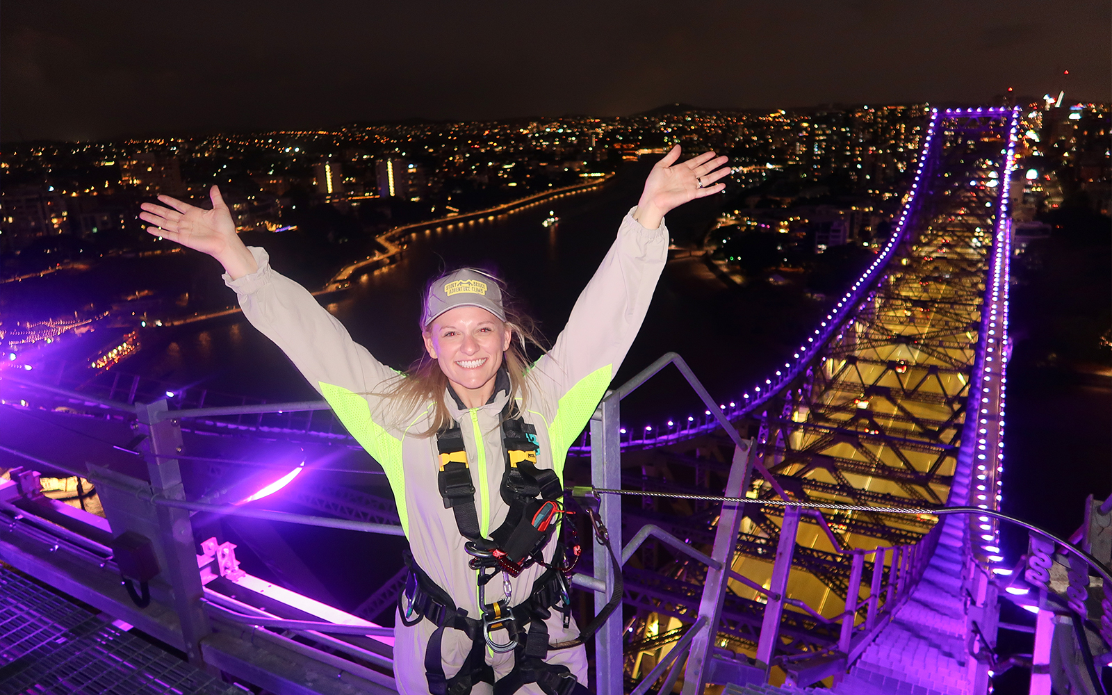 Person celebrating atop illuminated Story Bridge during night climb in Brisbane.