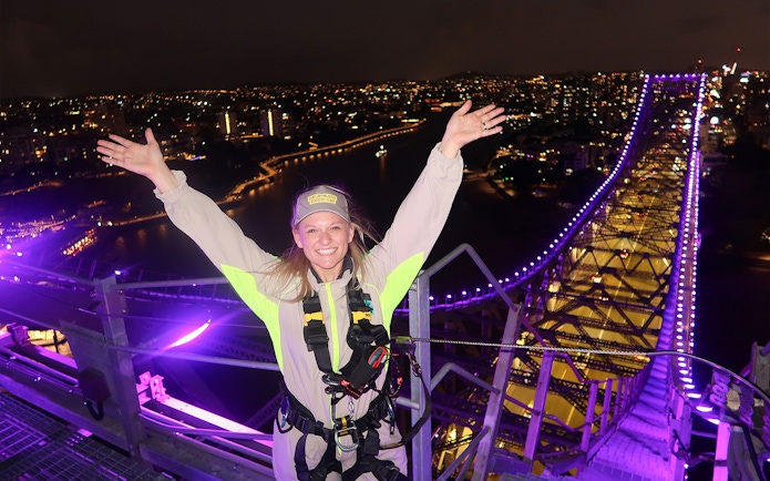 Person celebrating atop illuminated Story Bridge during night climb in Brisbane.