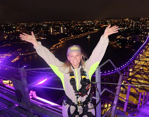 Person celebrating atop illuminated Story Bridge during night climb in Brisbane.