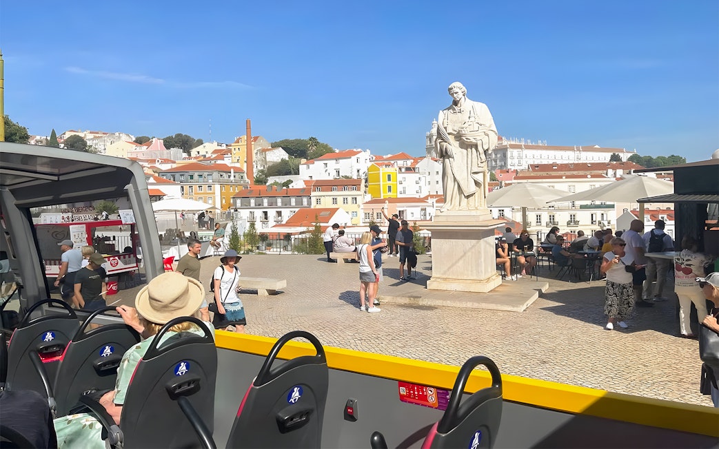Open-top bus view of Lisbon's Alfama district with a statue and tourists nearby.