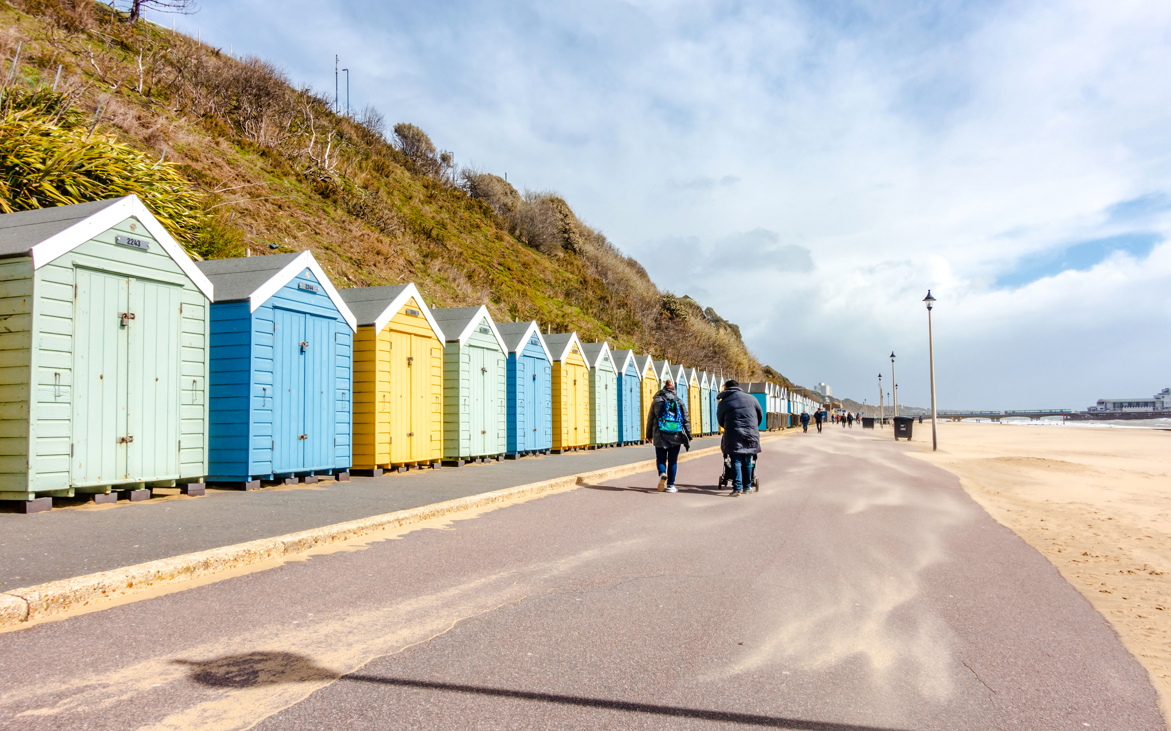 People walking along beach huts at Bournemouth Beach on a sunny day.