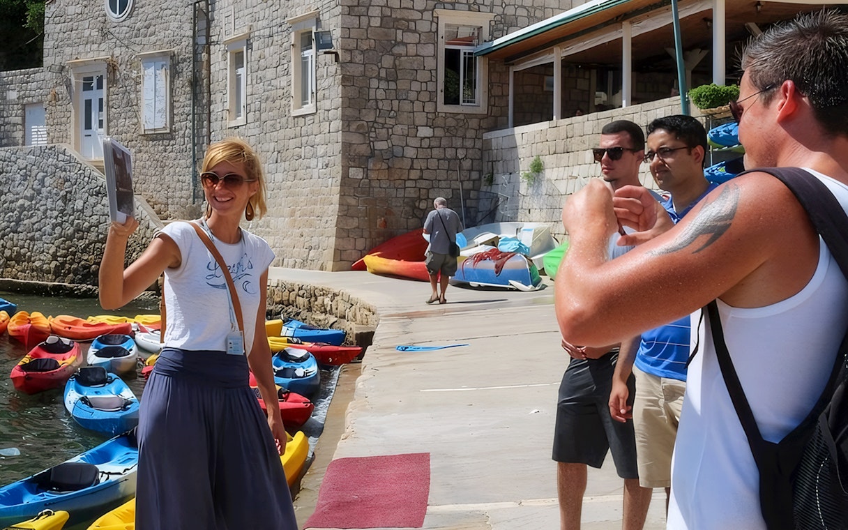 Female guide showing Game of Thrones location to tourists near kayaks and stone building.