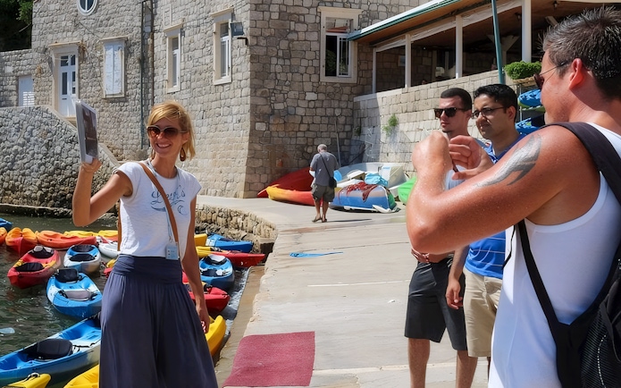 Female guide showing Game of Thrones location to tourists near kayaks and stone building.