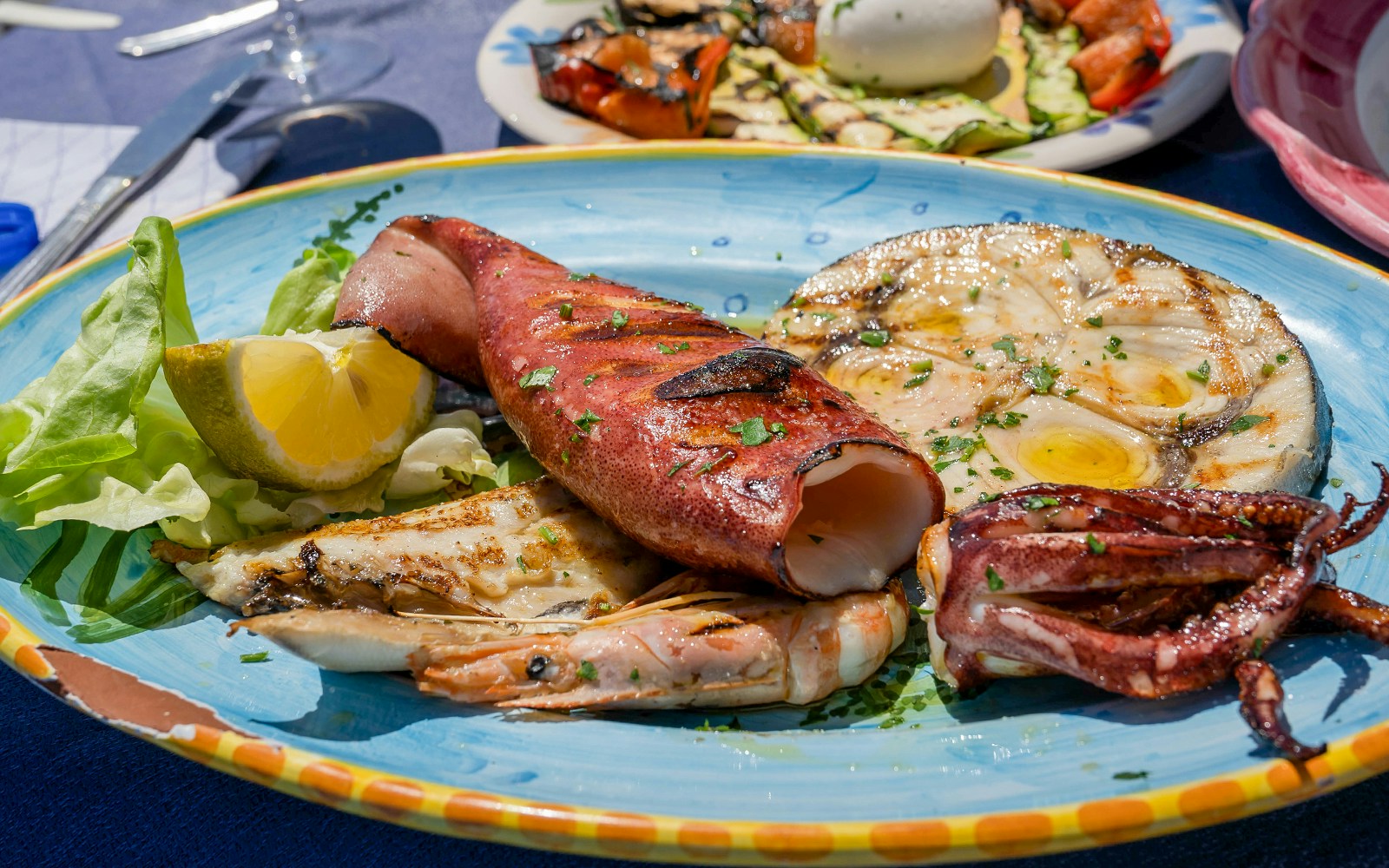 Grilled fish and octopus served with lemon and greens on a blue plate.