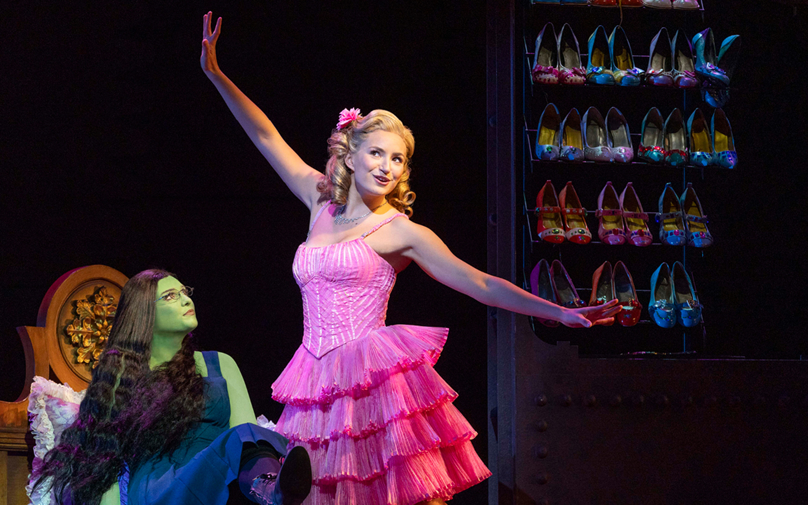 Two characters from the musical Wicked, one in a pink dress, with a shoe rack in the background.