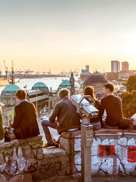 People sitting on a stone wall overlooking Hamburg harbor at sunset, with a telescope nearby.
