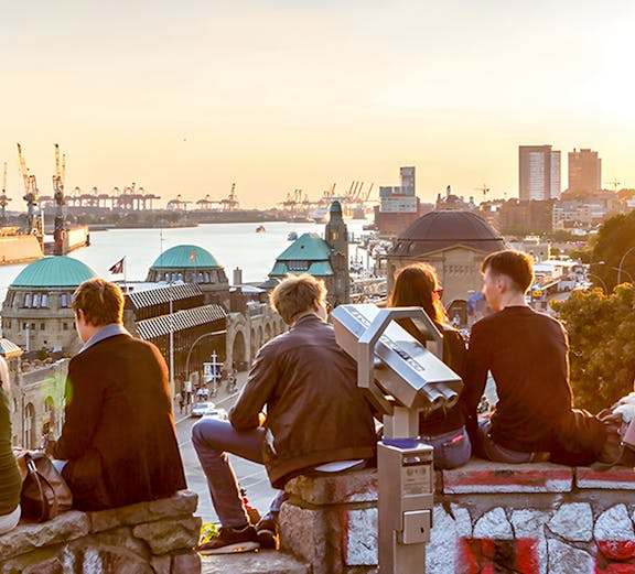 People sitting on a stone wall overlooking Hamburg harbor at sunset, with a telescope nearby.