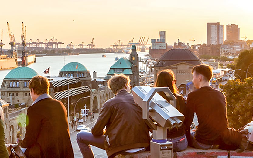 People sitting on a stone wall overlooking Hamburg harbor at sunset, with a telescope nearby.