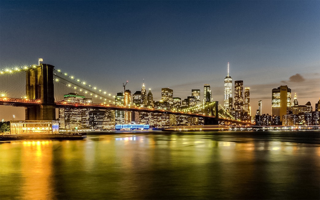 Brooklyn Bridge and Manhattan skyline illuminated at sunset during Statue of Liberty & Ellis Island cruise.