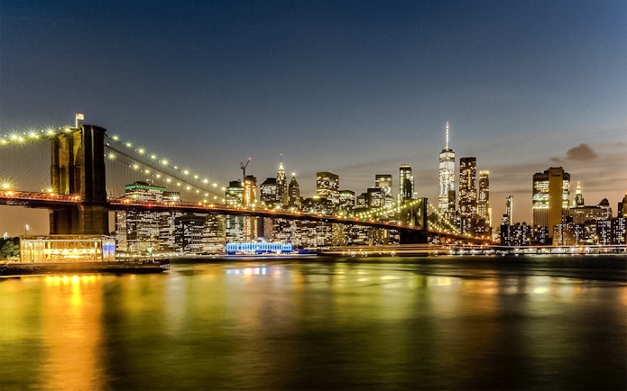 Brooklyn Bridge and Manhattan skyline illuminated at sunset during Statue of Liberty & Ellis Island cruise.