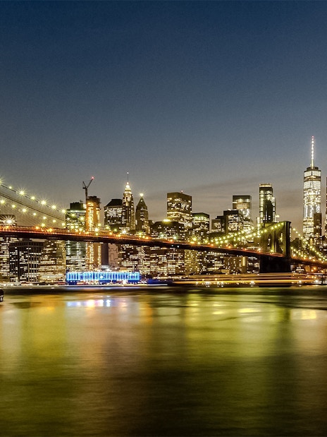 Brooklyn Bridge and Manhattan skyline illuminated at sunset during Statue of Liberty & Ellis Island cruise.