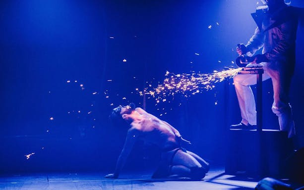 Performer kneeling on stage with sparks flying from a grinder in a dramatic Bite Club show.
