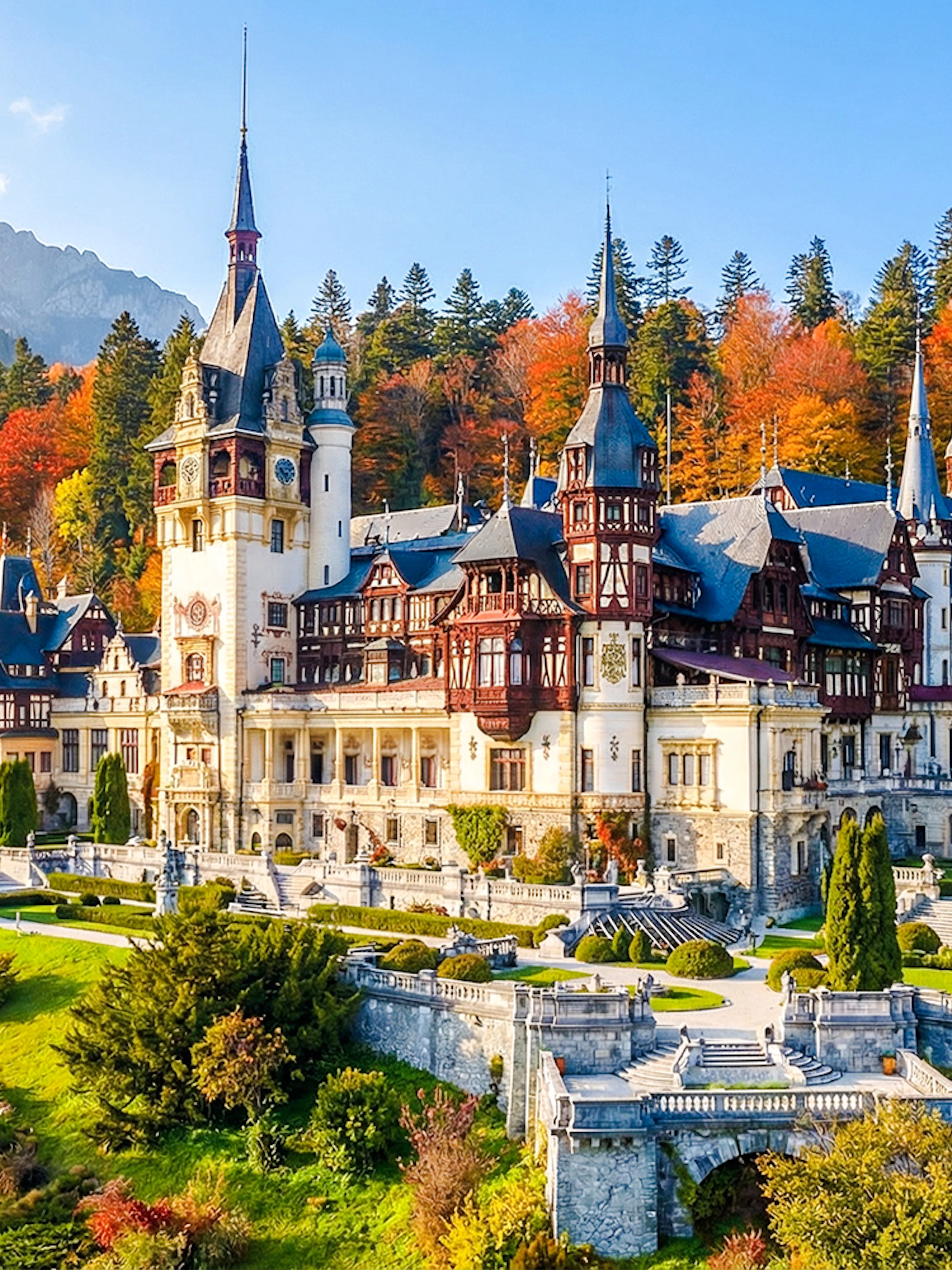Peleș Castle in Romania surrounded by autumn trees and mountains.