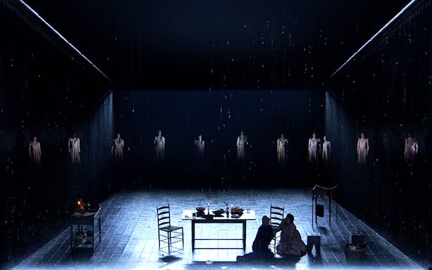 Stage scene from "The Crucible" with actors in dim lighting and rain effect.