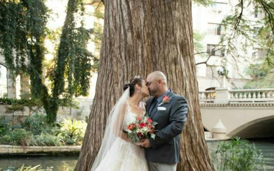Couple embracing by a tree near San Antonio River Walk bridge, part of self-guided city exploration game.