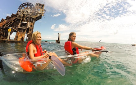 Two women kayaking in transparent kayaks near a rusted shipwreck in clear ocean waters.