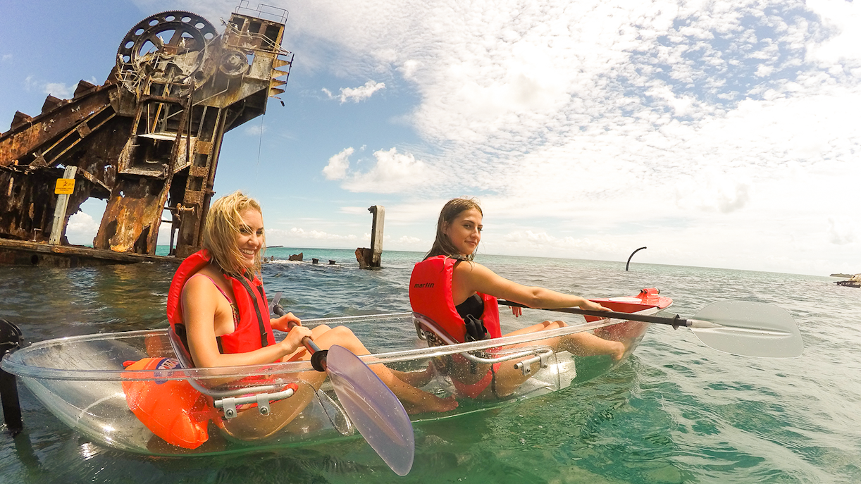 Two women kayaking in transparent kayaks near a rusted shipwreck in clear ocean waters.
