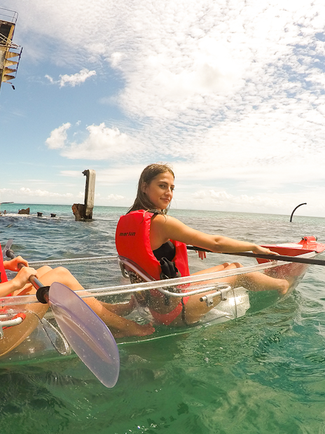 Two women kayaking in transparent kayaks near a rusted shipwreck in clear ocean waters.
