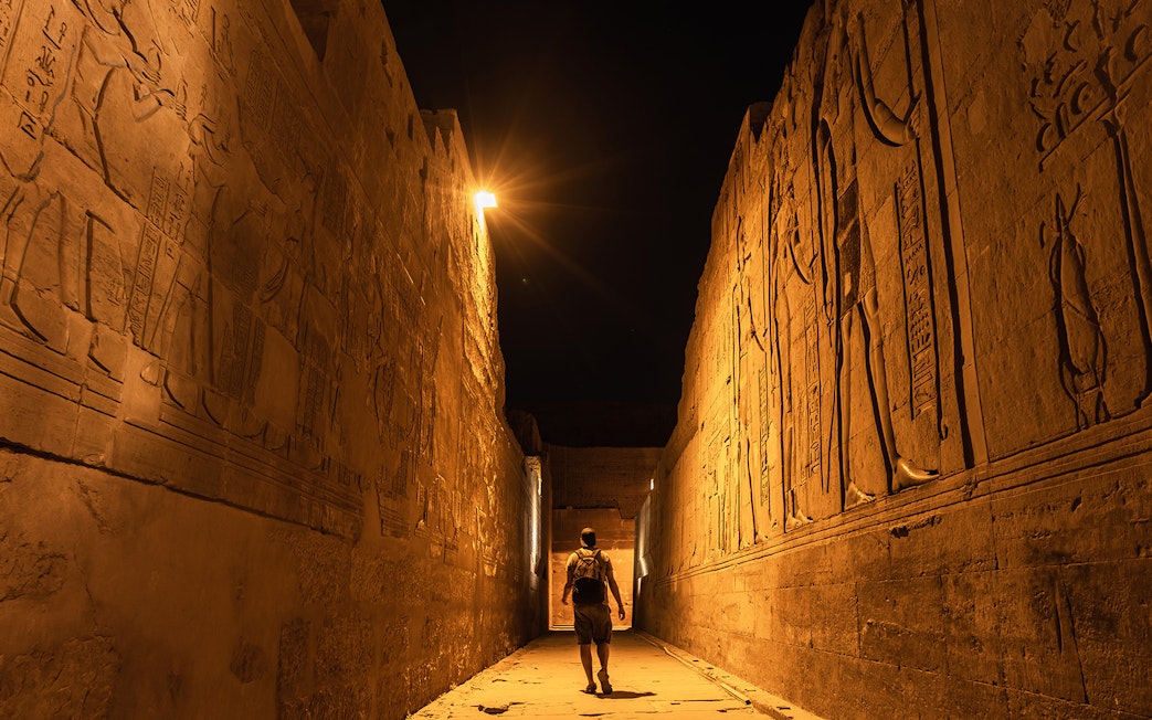 Traveler walking through illuminated ancient Egyptian temple corridor at night, Aswan to Luxor Nile cruise.