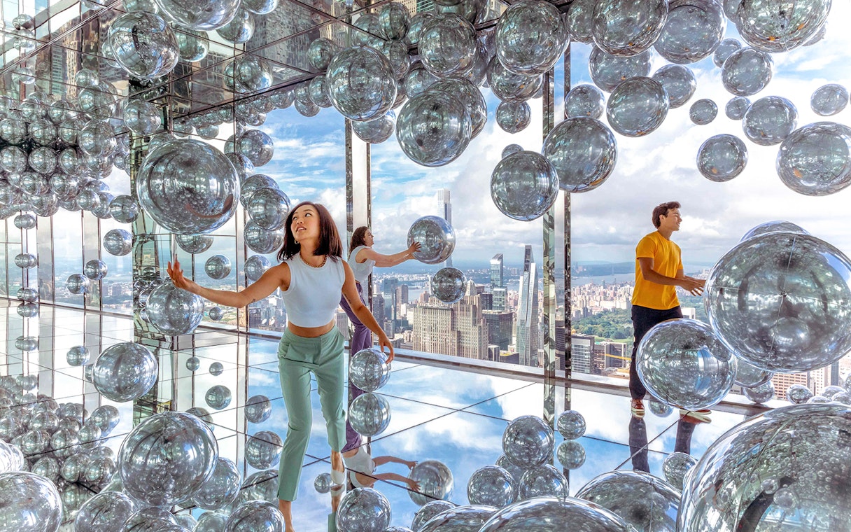 Visitors exploring mirrored room with reflective spheres, New York City skyline in background.