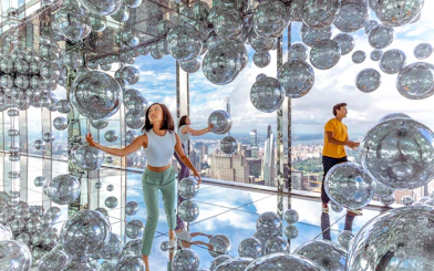 Visitors exploring mirrored room with reflective spheres, New York City skyline in background.