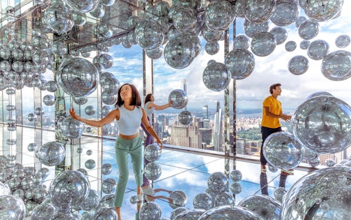Visitors exploring mirrored room with reflective spheres, New York City skyline in background.