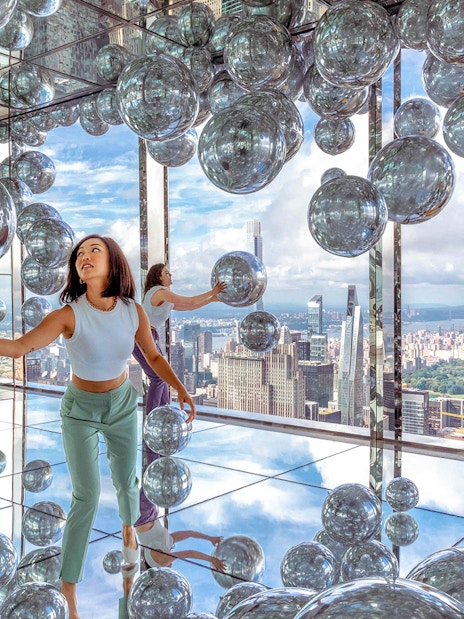 Visitors exploring mirrored room with reflective spheres, New York City skyline in background.