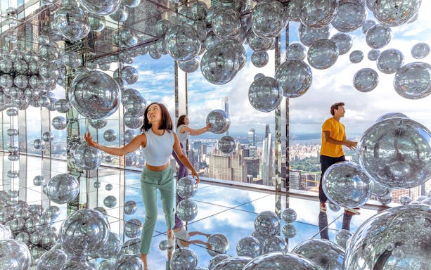 Visitors exploring mirrored room with reflective spheres, New York City skyline in background.