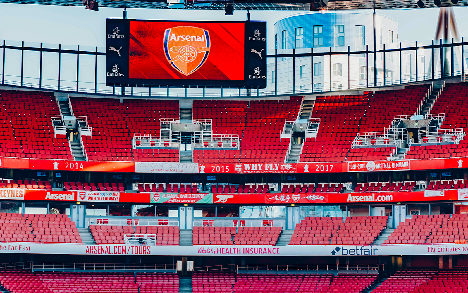 Emirates Stadium interior with red seats and Arsenal logo on screen.