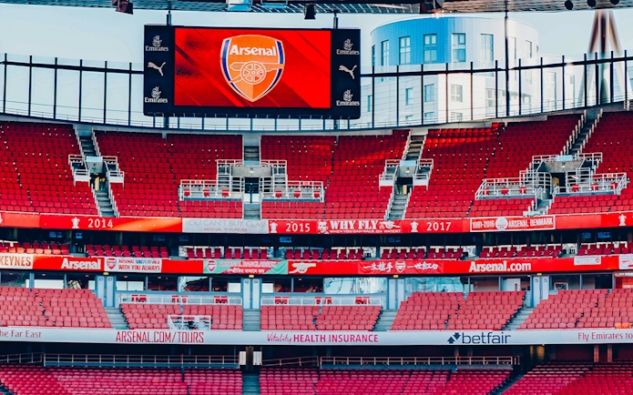 Emirates Stadium interior with red seats and Arsenal logo on screen.