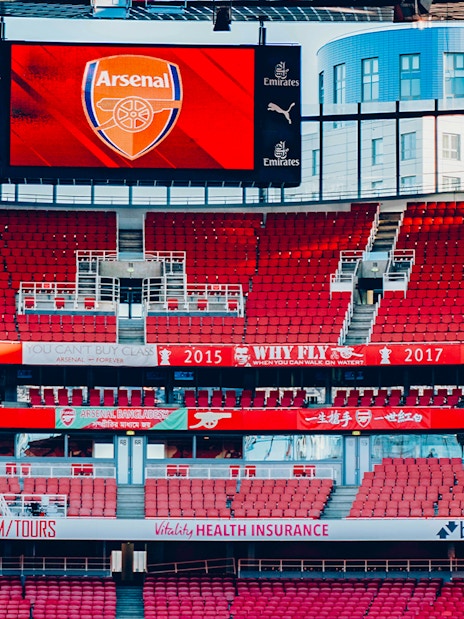 Emirates Stadium interior with red seats and Arsenal logo on screen.