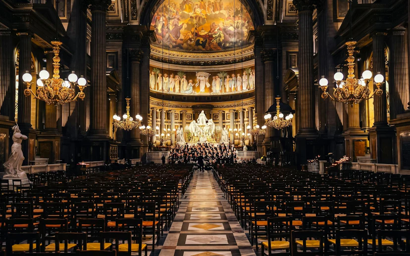Orchestra Hélios performing in Église de la Madeleine, Paris, with ornate chandeliers and frescoes.