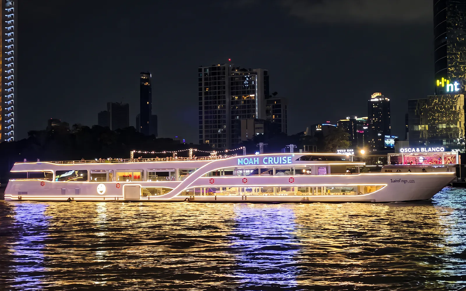 Noah Cruise ship illuminated at night on the Chao Phraya River, Bangkok skyline in background.