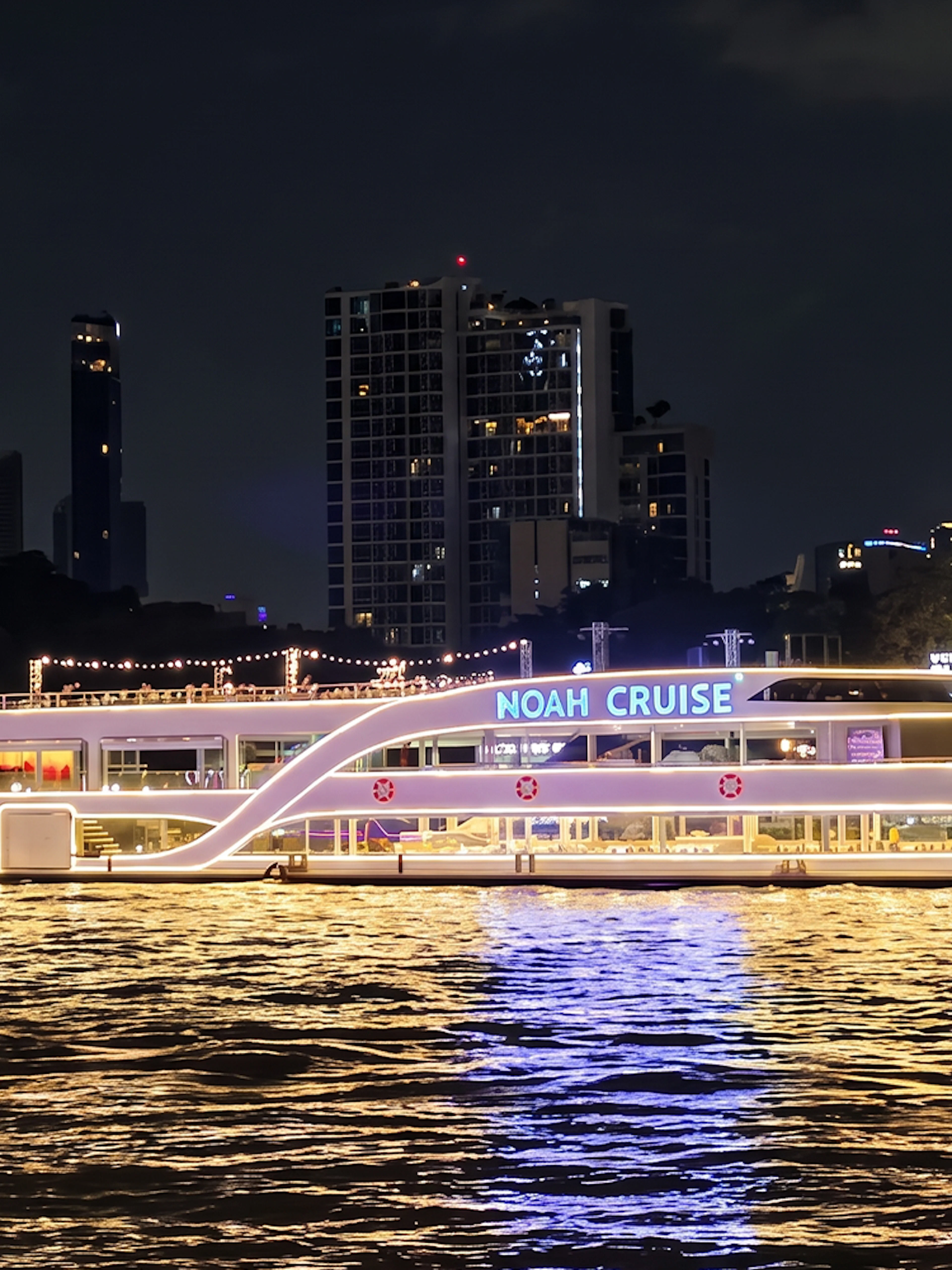 Noah Cruise ship illuminated at night on the Chao Phraya River, Bangkok skyline in background.