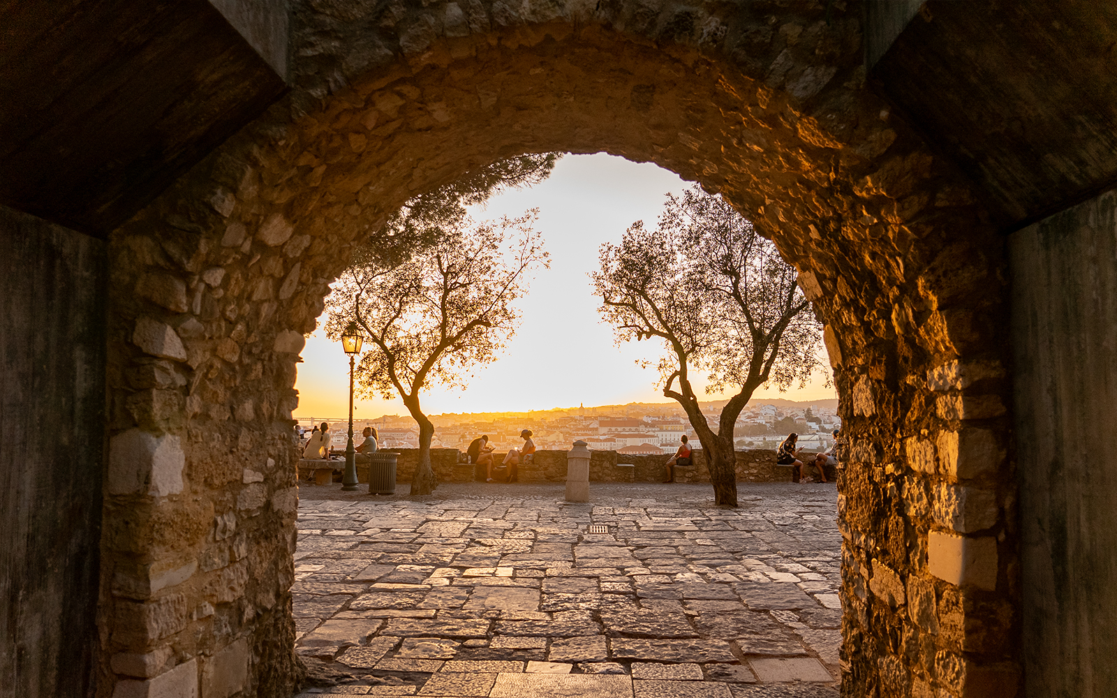 Archway view of sunset at St. George’s Castle, Lisbon, with people sitting near trees.