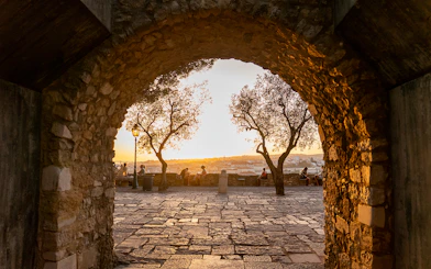 Archway view of sunset at St. George’s Castle, Lisbon, with people sitting near trees.