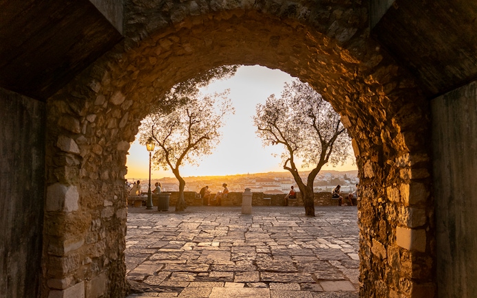 Archway view of sunset at St. George’s Castle, Lisbon, with people sitting near trees.