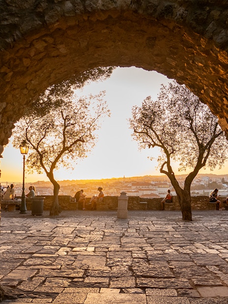Archway view of sunset at St. George’s Castle, Lisbon, with people sitting near trees.