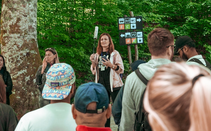 Group listening to a guide in a forested area with directional signs.