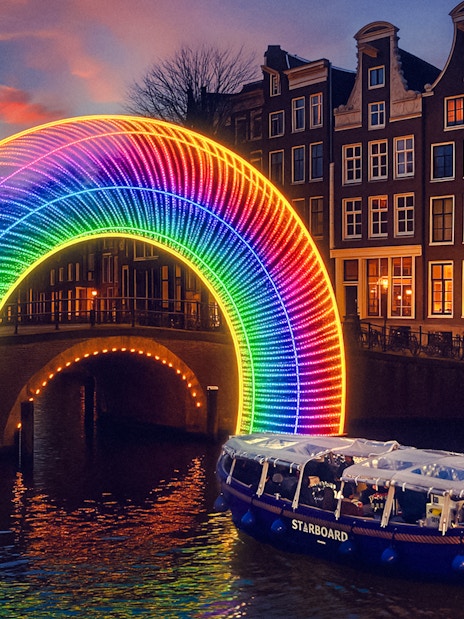 Amsterdam canal cruise passing under illuminated rainbow arch during Light Festival.