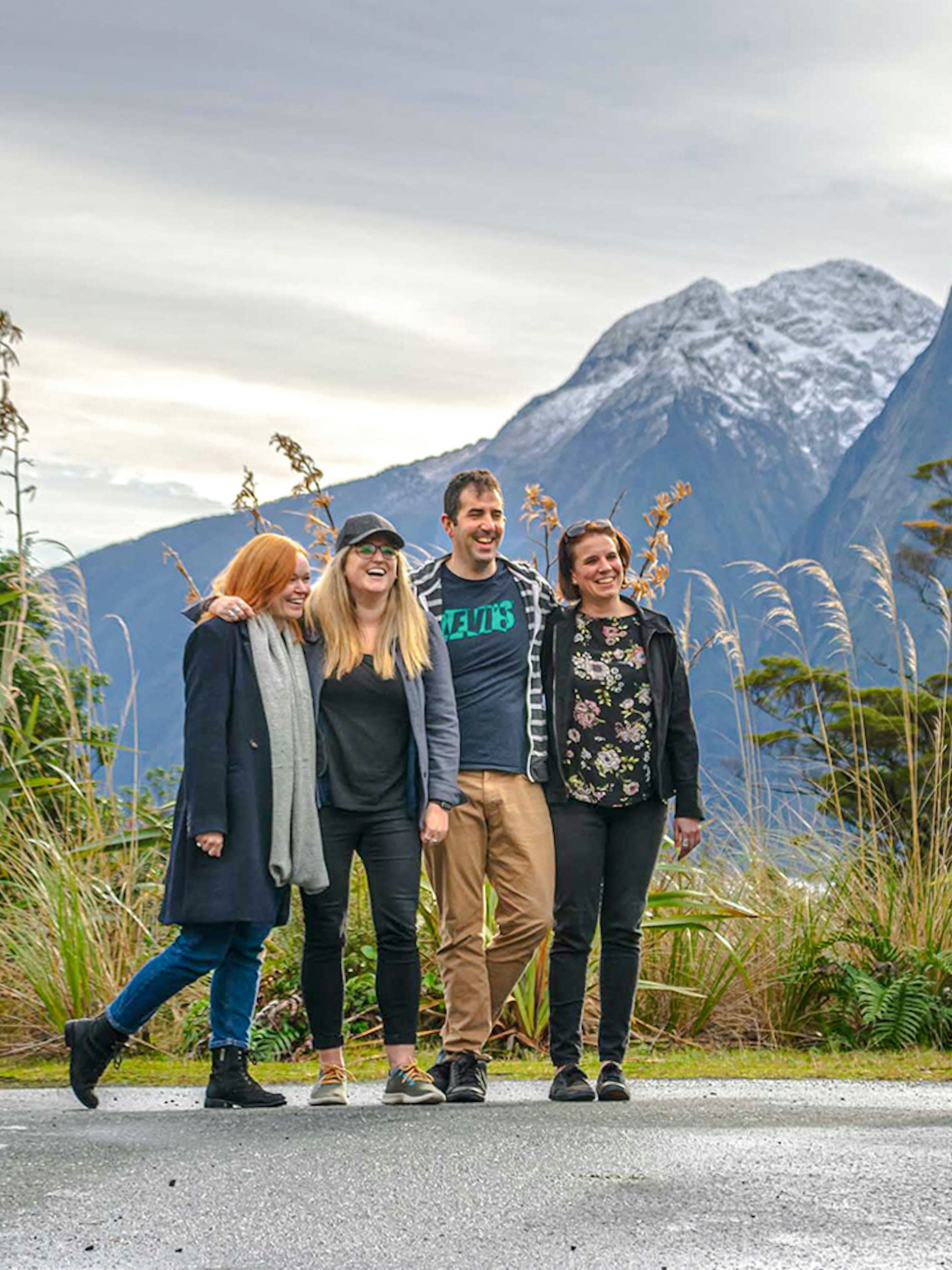 Group posing for a photo with a tour bus and snow-capped mountains in Milford Sound, New Zealand.