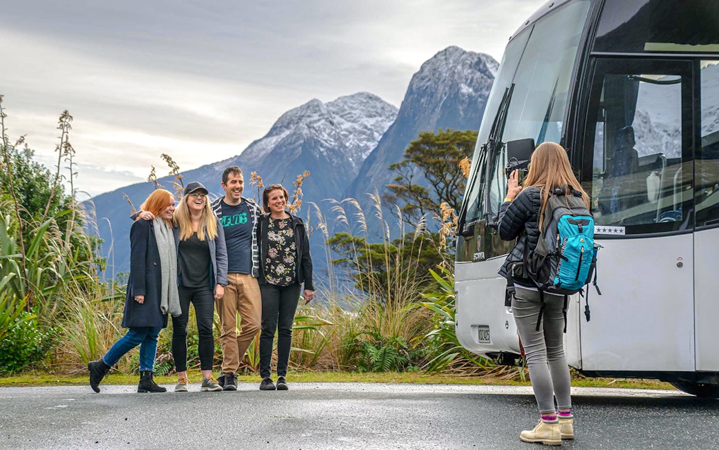 Group posing for a photo with a tour bus and snow-capped mountains in Milford Sound, New Zealand.