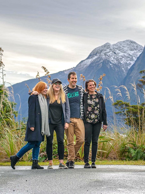 Group posing for a photo with a tour bus and snow-capped mountains in Milford Sound, New Zealand.